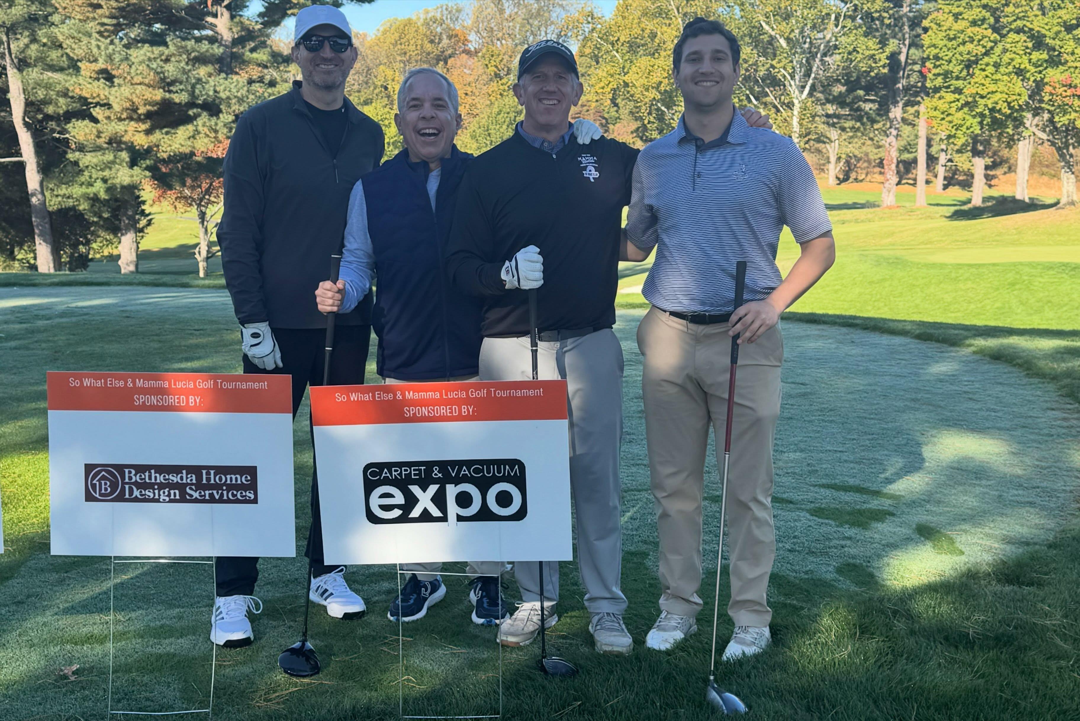 Golfers beside Carpet & Vacuum Expo and Bethesda Home Design Services sponsor signs at the So What Else and Mamma Lucia tournament
